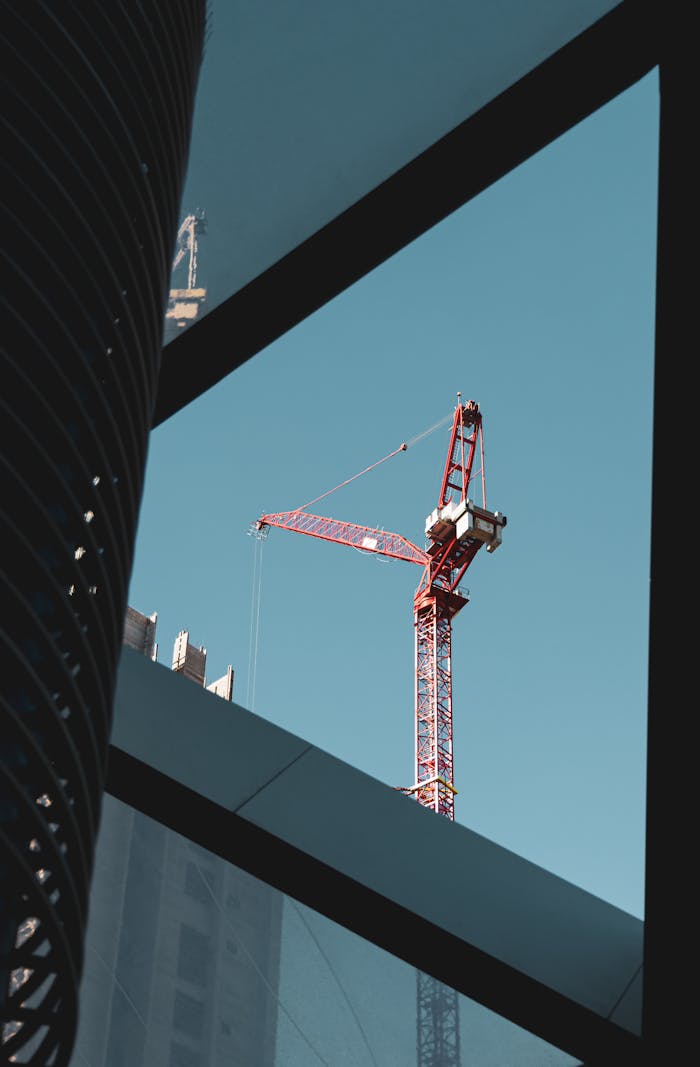 why-choose-us A red tower crane in a city construction setting framed by modern architecture, clear blue sky.