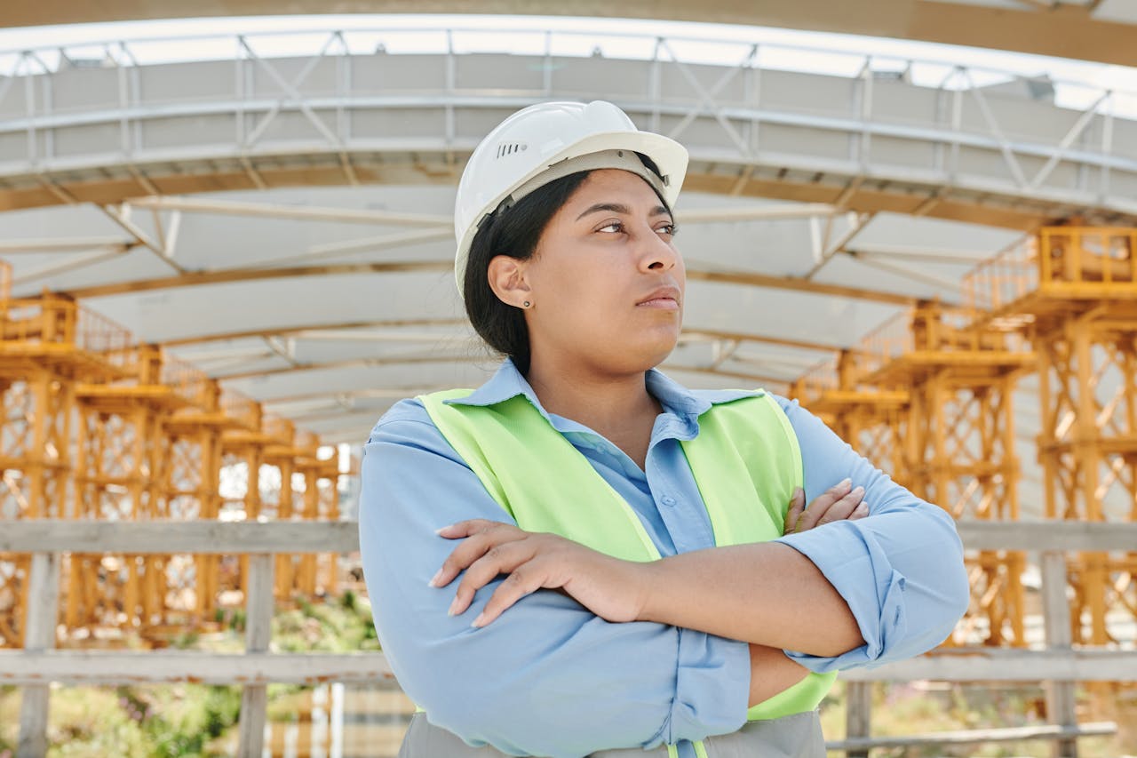gallery-4 Young female engineer crossing her arms at a construction site, wearing a hardhat and reflective vest.